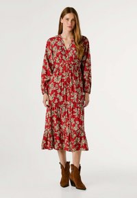 Robe maxi en tissu rouge avec un motif floral crème, décolleté en v, manches longues et jupe à volants. Portée avec des bottes cheville marron.
