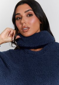 Maglione in pile blu navy con colletto alto e ripiegato. Tessuto morbido e testurizzato con una vestibilità ampia. Presenta un collo rotondo e maniche larghe.