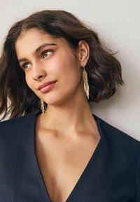 Goldfarbene, hängende Ohrringe mit einem strukturierten Kettendesign und scharfen, länglichen Akzenten. Getragen von einem Model mit schulterlangem Haar.