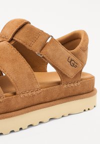 Brune semskede sandaler med åpen tå-design, justerbar reim og en teksturert lys tau gummisåle med lette spor. UGG-logo på siden.