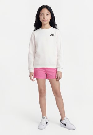 Meisje staand, gekleed in een wit Nike-sweatshirt, roze korte broek en witte Nike-sneakers tegen een effen witte achtergrond.