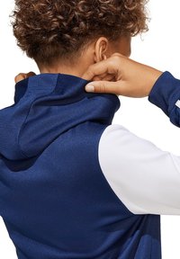 Navyblauwe hoodie van een textuurstof. Heeft een hoge kraag en witte mouwen. Hand die de kraag vastgrijpt, toont het zachte materiaal.