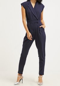 Marinblå jumpsuit med blazermodell, korta ärmar, bälte i midjan och sidofickor, matchad med silverfärgade högklackade skor.