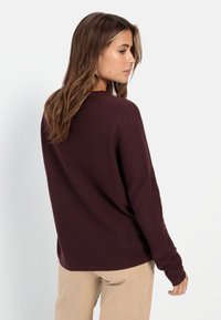 Burgunderfarbener Strickpullover mit einem runden Halsausschnitt und langen Ärmeln, Merkmale sind der lockere Schnitt und die gerippte Textur. Sichtbare Nähte an den Schultern.