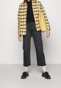 Camicia di flanella gialla e nera a quadri, maglione nero, jeans scuri e scarpe nere con frange. Borsa gialla. Sfondo semplice.