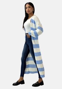 Lång stickad kofta i blå och vita ränder, kombinerad med en åtsittande vit topp och mörka jeans. Blanka svarta loafers fulländar looken.
