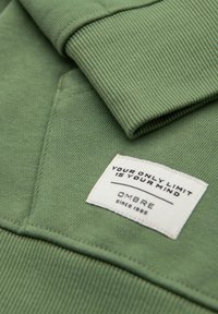 Grøn sweatshirtstof med ribbede håndled, der har et vævet mærke med sort tekst, der siger: "DIN ENESTE GRÆNSE ER DIN HJERNE," samt branddetaljer.