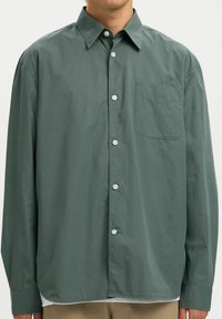 Homme portant une chemise boutonnée à manches longues de couleur vert foncé, ample, avec une poche poitrine avant, portée sur un pantalon beige.