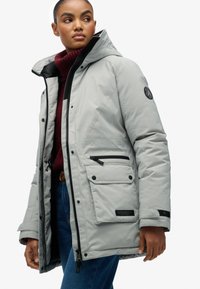 Kobieta w jasnoszarej zimowej kurtce z kapturem, nosząca bordowy golf i niebieskie dżinsy, patrząca w bok.