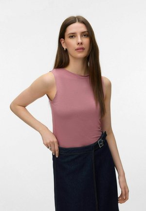 Woman with long brown hair wearing a sleeveless mauve top and dark denim skirt with a side buckle, standing against a plain background.