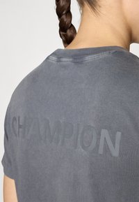 T-shirt en coton gris avec un grand logo "CHAMPION" en relief, discret, au dos. Manches courtes et design à col rond.