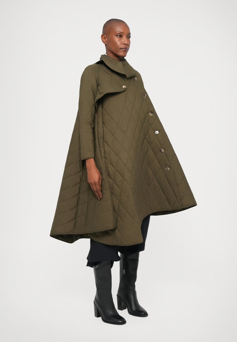 Femme portant un manteau en cape matelassé vert olive avec des boutons, associé à un pantalon noir et des bottes noires à talons montant jusqu'au genou, se tenant devant un fond blanc.