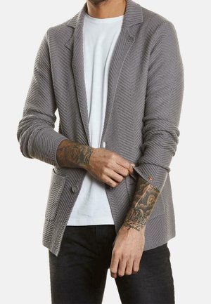Hombre con blazer de punto gris texturizado sobre camiseta blanca, remangándose una manga, mostrando antebrazos y muñecas tatuados.