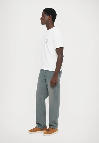 Vit kortärmad T-shirt med en liten svart smiley-logotyp på bröstet, i kombination med lösa grå denimjeans och bruna mockaskor.