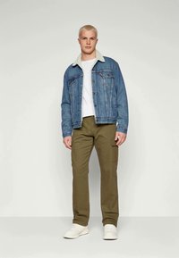 Jeansjacke mit Lammfellkragen, kombiniert mit olivgrünen Cargo-Hosen. Darunter ein weißes T-Shirt und weiße Sneaker, stehend vor einem neutralen Hintergrund.