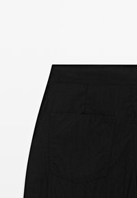 Vue arrière d'un pantalon noir montrant une ceinture plate et une grande poche plaquée sur le côté droit.