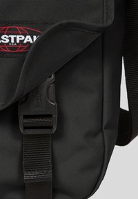Sac à bandoulière noir avec une texture lisse, doté d'un rabat maintenu par une boucle en plastique, et arborant un logo rouge et blanc bien en vue.