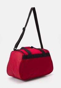 Bolsa de deportes roja hecha de un tejido duradero con asas negras. Forma redondeada, parte superior con cremallera y base plana para estabilidad.