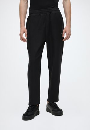 Mand iført sorte slim fit joggingbukser med elastisk talje og sorte platform sneakers, der står på hvid baggrund.