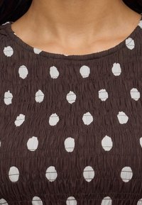 Personne portant un haut marron texturé avec des pois blancs irréguliers, montrant le décolleté et la partie supérieure de la poitrine.