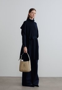 Ναυτικό μπλε πλεκτό τούνικ με ζώνη στη μέση, συνδυασμένο με σκούρο παντελόνι φαρδιάς γραμμής. Κρατάει μια δομημένη μπεζ τσάντα χειρός με λογότυπο στο μπροστινό μέρος.