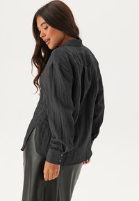 Camicia nera a maniche lunghe con finitura testurizzata, dotata di colletto button-down e polsini. La camicia ha una vestibilità rilassata e spacchi laterali.