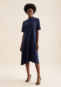 Abito midi blu navy con maniche corte, colletto rigido e orlo asimmetrico. Tessuto leggero e design minimalista senza motivi.