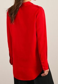 Rode blouse met lange mouwen, gladde textuur, afgeronde zoom, met knopen aan de mouwen en een kraag. Beschikt over een achterpand met inzetten.