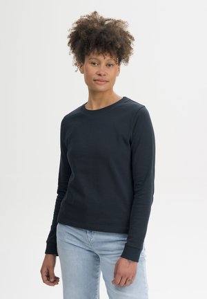 Vrouw met krullend haar die een zwart shirt met lange mouwen en lichtblauwe jeans draagt, staand tegen een effen witte achtergrond.