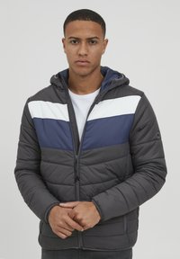 Veste rembourrée grise avec des rayures horizontales bleu marine et blanches. Comprend une capuche, une fermeture éclair à l'avant et des poignets élastiques. Texture lisse, design décontracté.