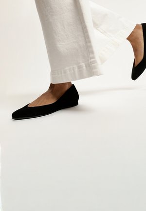 Ballerine nere in suede con design a punta e tacco basso, indossate con pantaloni bianchi a gamba larga, che mettono in evidenza la silhouette della scarpa.