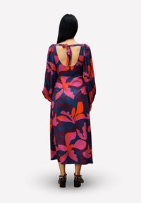 Eine Frau mit langen schwarzen Haaren steht mit dem Rücken zur Kamera. Sie trägt ein langärmeliges Midi-Kleid mit großen rosa und orangefarbenen Blumenmustern sowie einem offenen Rücken mit Bindeband.