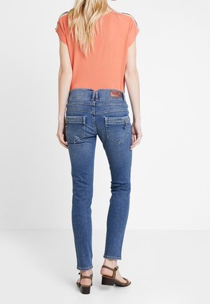 Kvinde iført stramme blå jeans, koral-farvet kortærmet top og brune sandaletter med hæl, stående med ryggen mod seeren mod en ensfarvet baggrund.