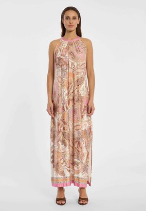 Frau trägt ein ärmelloses, bodenlanges Kleid mit Paisley-Muster in Pink-, Orange- und Beigetönen, kombiniert mit braunen Sandalen mit offenen Zehen, steht vor weißem Hintergrund.