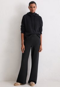 Czarna, oversize'owa bluza z kapturem i sznurkami, w zestawieniu z czarnymi, szerokimi spodniami w prążki oraz lekkimi brązowymi sneakersami.