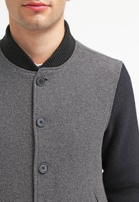 Grijze wolblend jas met zwarte mouwen, ronde kraag en een vijf-knopenfront. Heeft een gestructureerde stof en een geribbelde kraag voor extra detail.