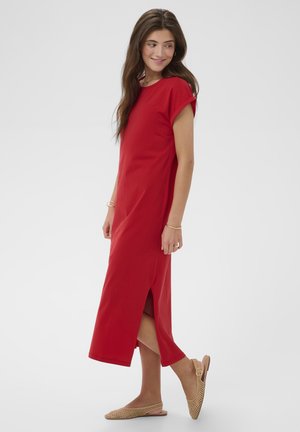 Frau mit langen braunen Haaren, die ein rotes Midikleid mit seitlichem Schlitz und beigefarbene gewebte Slingback-Ballerinas trägt, steht vor einem schlichten Hintergrund.