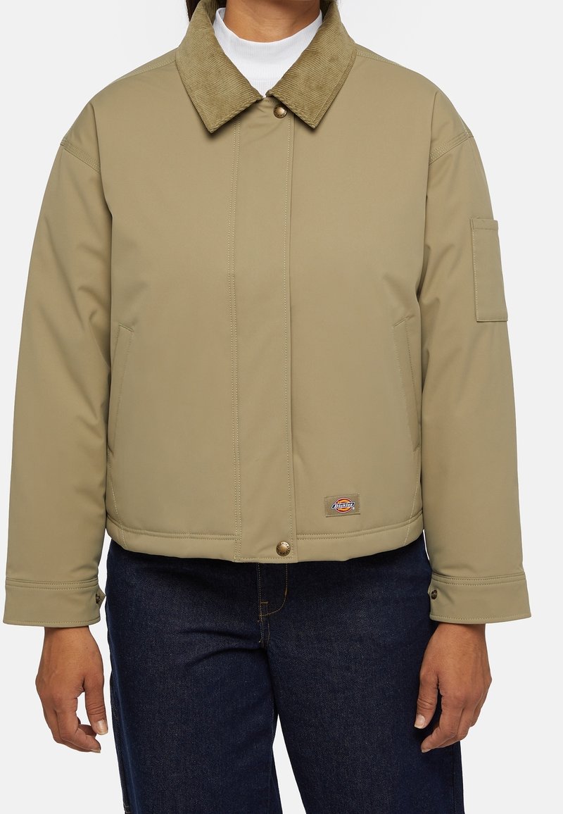 Personne portant une veste beige avec un col en velours côtelé, des boutons-pression, des poches latérales et un jean bleu foncé.