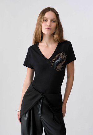 Femme aux longs cheveux bruns portant un t-shirt noir à col en V orné de détails métalliques et un pantalon noir drapé, debout devant un fond uni.