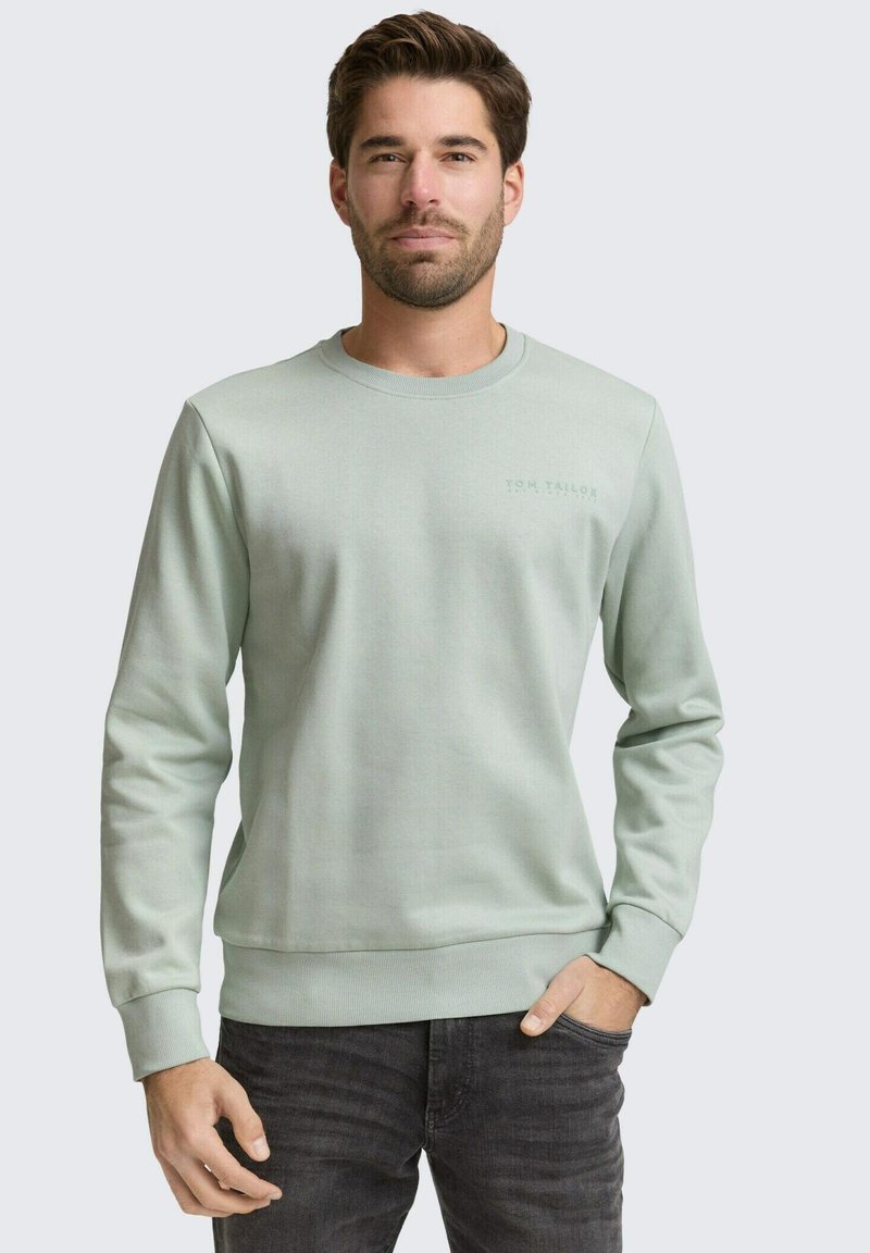 Homme portant un sweat-shirt à manches longues vert clair et un jean foncé, debout avec une main dans la poche devant un fond uni.