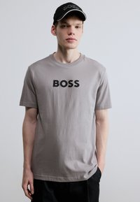 Γκρι βαμβακερό μπλουζάκι με την επιγραφή "BOSS" σε μαύρο. Κοντά μανίκια, στρογγυλή λαιμόκοψη, άνετη εφαρμογή. Μαύρο καπέλο με το ίδιο λογότυπο της μάρκας σε λευκό.