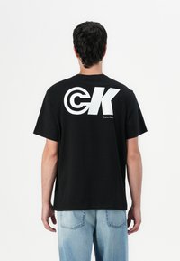 T-shirt de algodão preta com grande logótipo branco "CK" nas costas, com o texto "Calvin Klein" por baixo. Corte casual, mangas curtas, gola redonda.