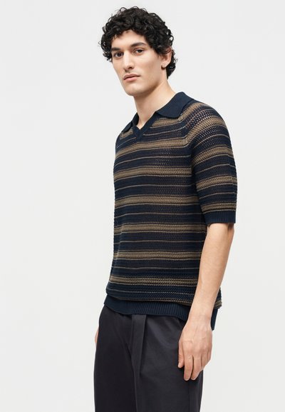 Jeune homme aux cheveux bouclés portant un polo tricoté à manches courtes rayé bleu marine et marron ainsi qu'un pantalon foncé, debout devant un fond uni.