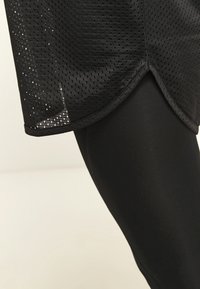 Czarny siatkowy koszulka sportowa z zaokrąglonym dołem, noszona na czarnych legginsach kompresyjnych. Posiada wentylowany materiał o gładkiej teksturze.