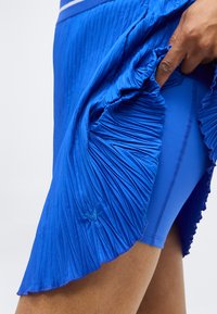 Χέρι σηκώνει πλισέ μπλε φούστα, αποκαλύπτοντας εφαρμοστά μπλε σορτς από κάτω, εστιάζοντας στην υφή και τη στρώση του αθλητικού ρουχισμού.