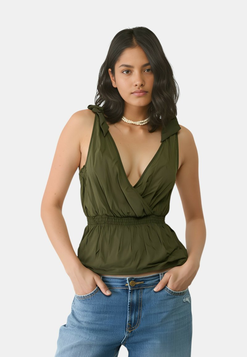 Top verde senza maniche con design avvolgente, tessuto leggero, vita arricciata e dettagli a fiocco sulle spalle; abbinato a jeans blu.