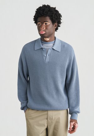 Jersey de punto acanalado azul claro con cuello con solapa y tres botones, usado sobre una camisa de manga larga a rayas. Corte informal.