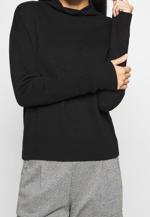 Frau trägt einen schwarzen, langärmeligen Strickpullover und graue, strukturierte Hosen, die Arme verschränkt und eine Hand berührt den anderen Arm.