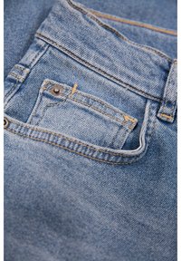 Jeans in denim con un lavaggio azzurro chiaro, caratterizzati da una tasca posteriore con cuciture visibili e un bottone di rame attraverso un piccolo lembo decorativo.