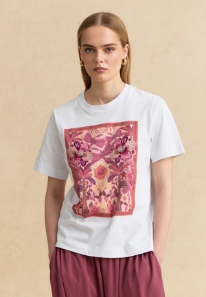 Donna che indossa una camicia bianca a maniche corte con grande motivo floreale rosa e uccelli, abbinata a pantaloni a vita alta color malva, sfondo neutro.